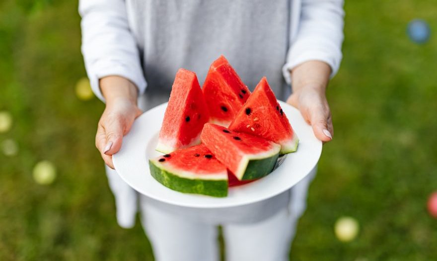 A woman is holding a white plate with five watermelon slices. She is outside, you can see the grass behind her.